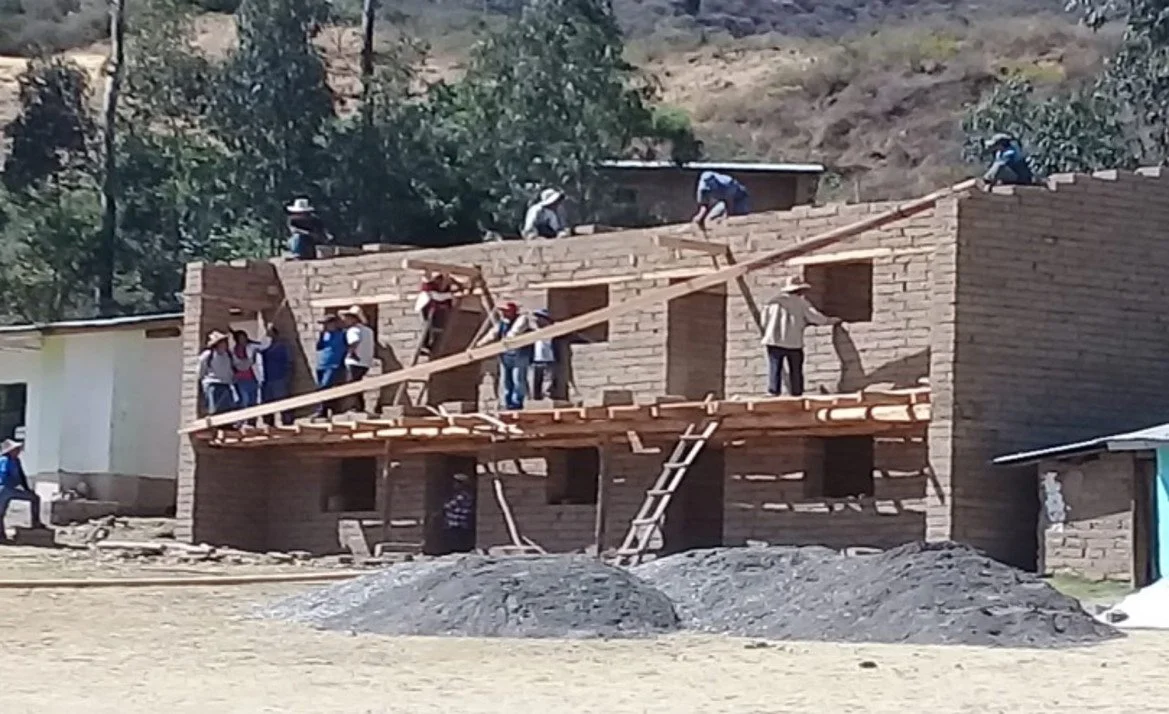 Grupo de personas trabajando en la construcción de una casa de ladrillos, algunos en el techo y otros en los pisos, con herramientas y materiales de construcción.