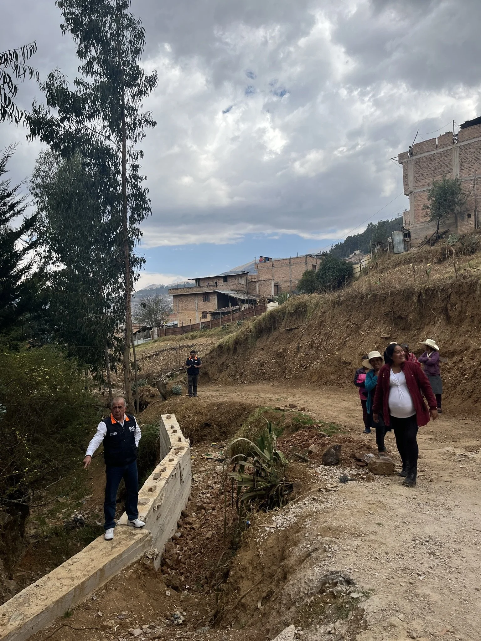 Grupo de personas caminando por un camino de tierra en una zona rural con casas de ladrillo en las colinas y árboles alrededor, bajo un cielo nublado.