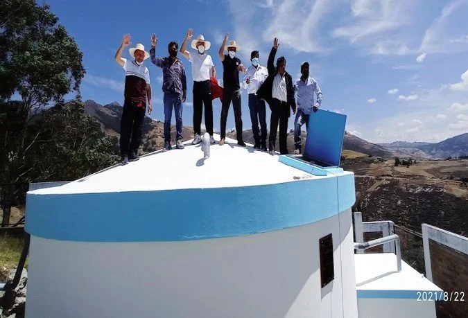 Grupo de ocho personas en la cima de una gran estructura con vista a un paisaje montañoso, algunos con sombrero y mascarilla, levantando las manos en señal de saludo o celebración.