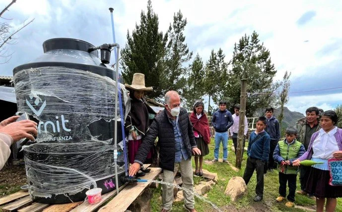 Varias personas, incluyendo adultos y niños, observan una demostración con un sistema de recolección de agua conectado a un gran tanque negro, en un entorno rural con árboles y césped.