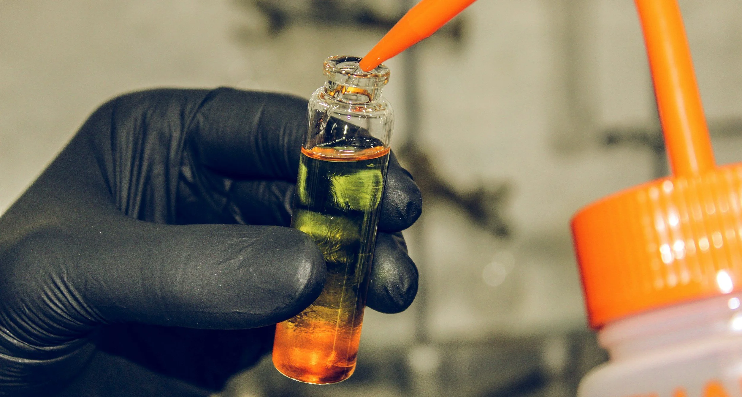 A gloved hand holding a small glass vial with a yellow-orange liquid inside, against a blurred background.