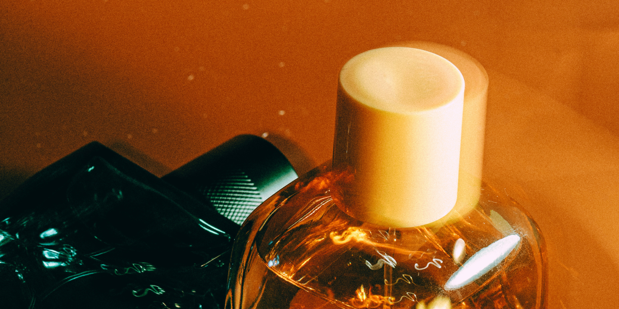 Close-up of perfume bottles, one with an amber liquid and a white cap, reflecting warm light, on a softly lit orange background.