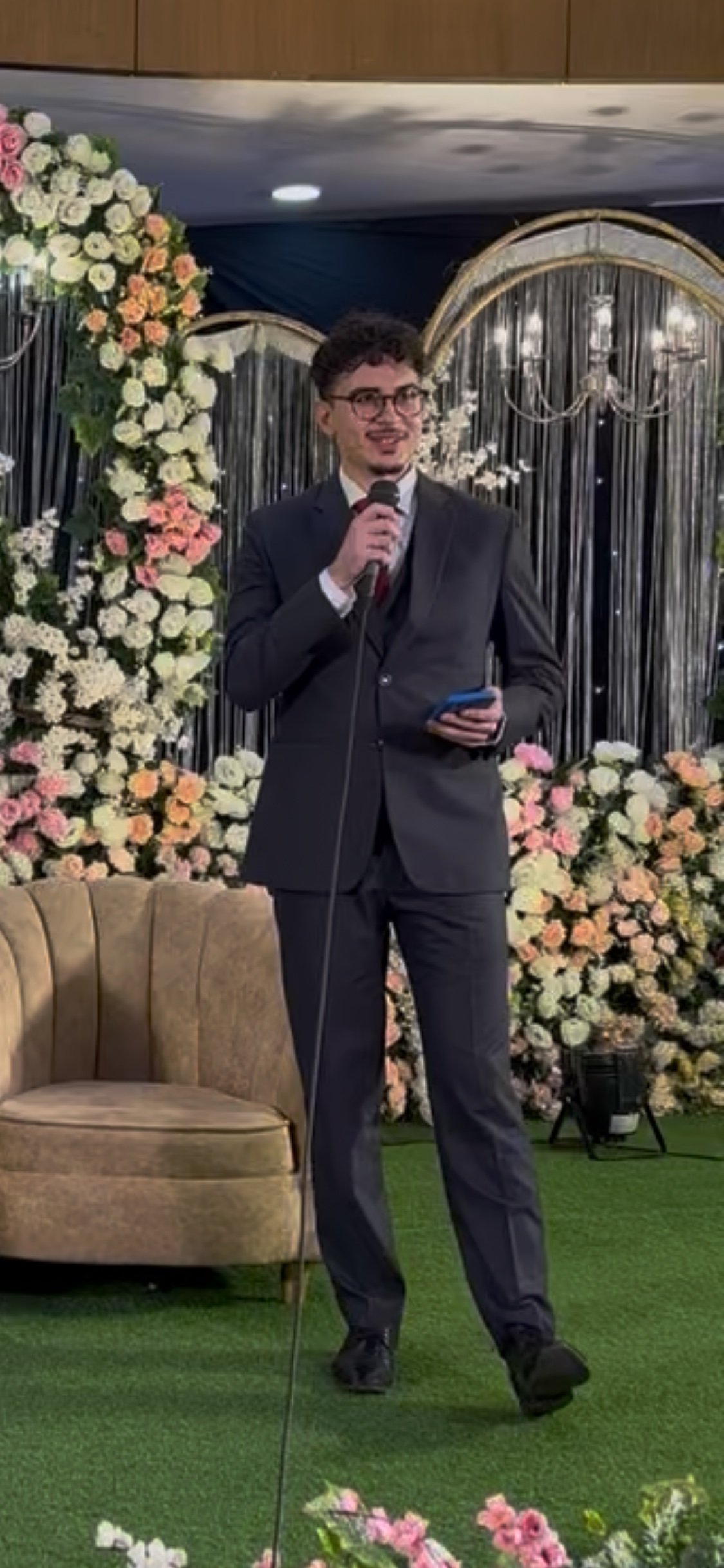 A man in a dark suit and glasses holding a microphone and a phone, standing in front of a floral backdrop at a wedding or event.