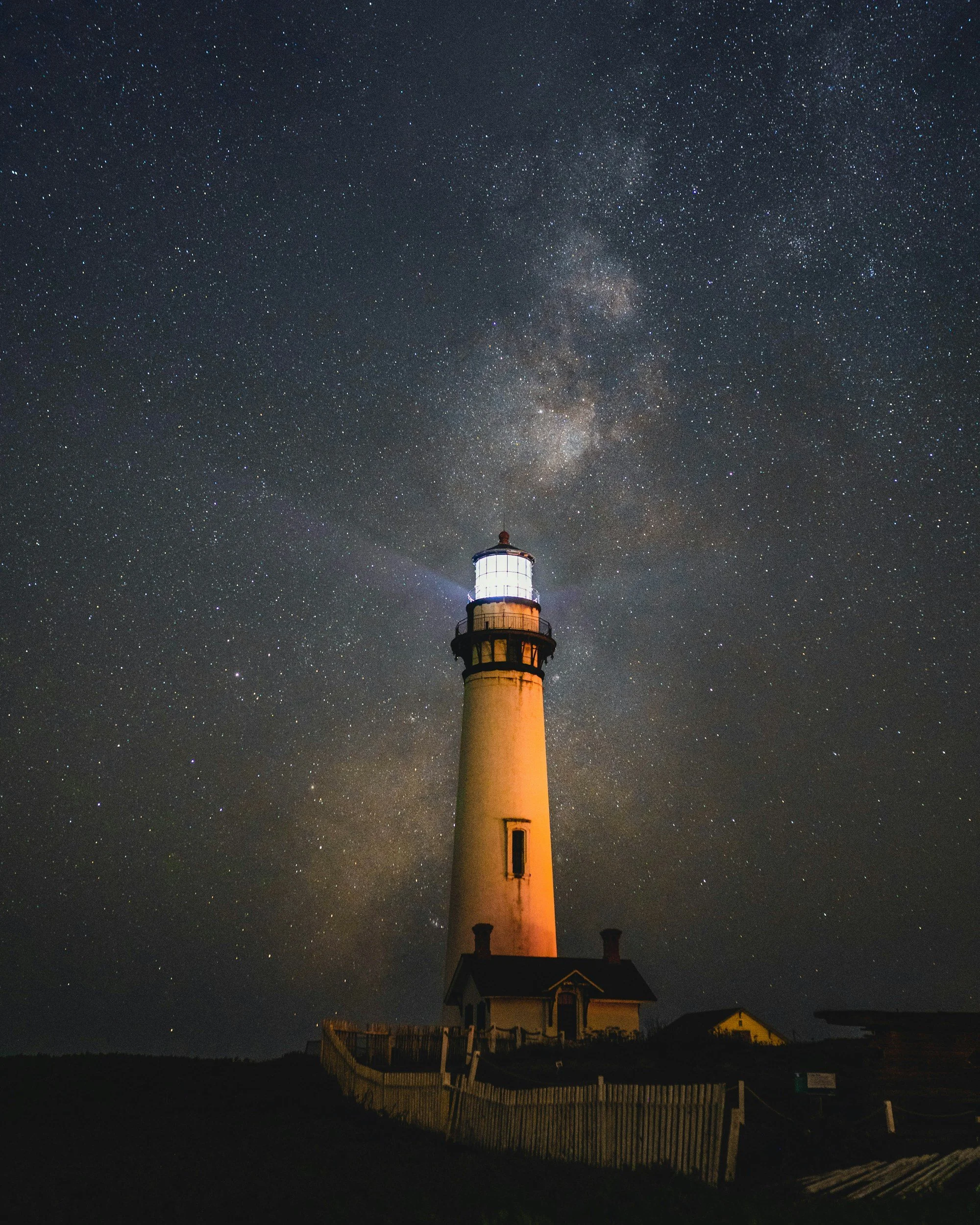 Nighttime scene featuring a lighthouse with a bright beacon and a star-filled sky, including the Milky Way galaxy.