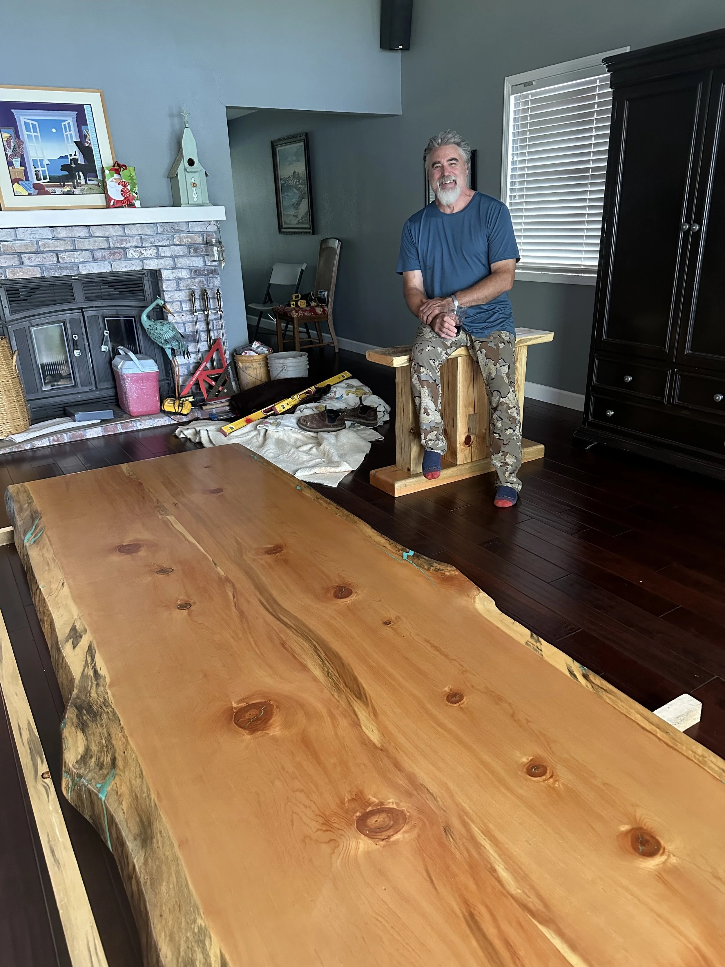 A man with a beard and gray hair, wearing a blue t-shirt and camouflage pants, smiling and sitting on a wooden block in a living room. The room has a brick fireplace, a large wooden table in the foreground, and various tools and materials on the floor, indicating woodworking or furniture-making activity.