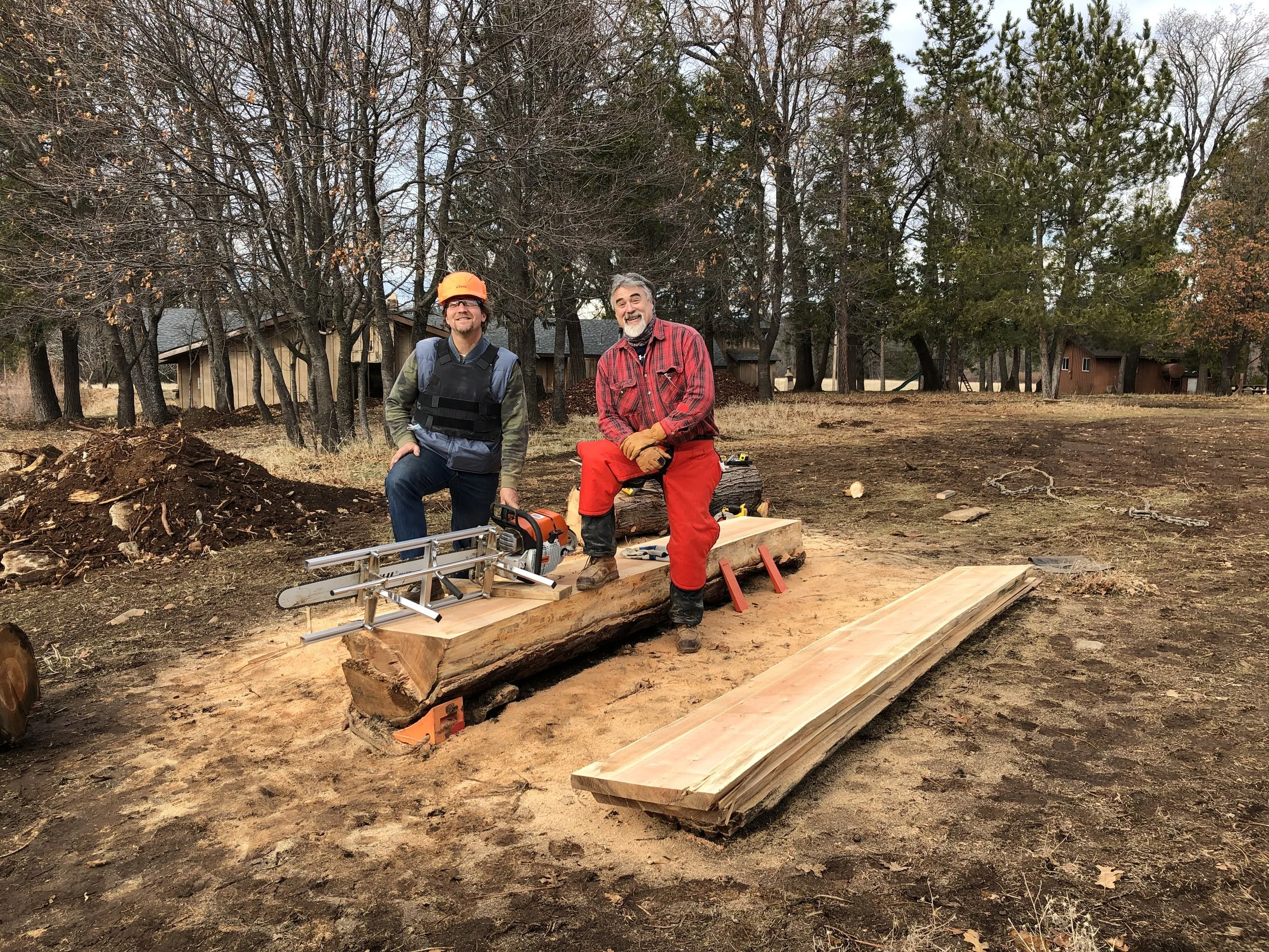 Two men working outdoors on a woodworking project, one with a chainsaw and the other sitting on a large log, with trees and houses in the background.
