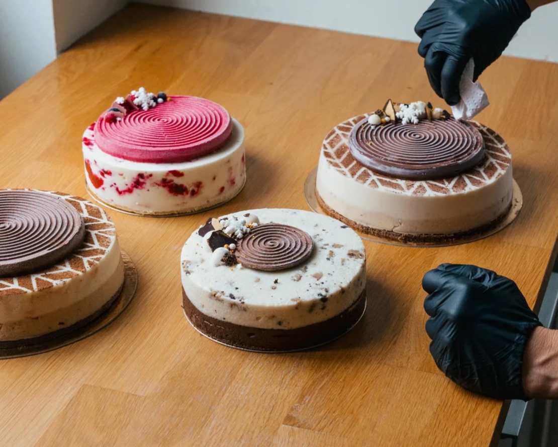 Vijf taarten op een houten tafel, met iemand die er een snijdt, allemaal versierd met chocolade en decoraties.