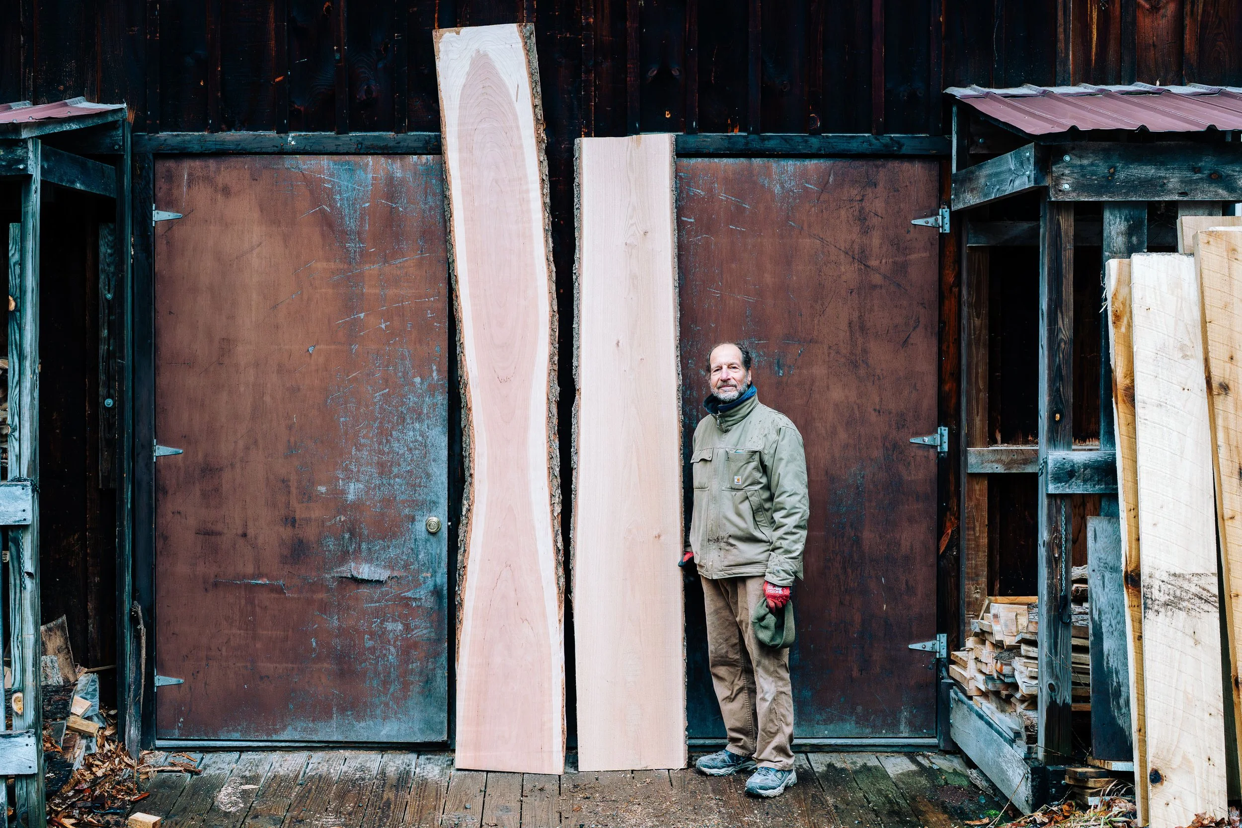 Mark stands next to two large live edge plank boards ready to become a beautiful bartop in front of the shop doors.