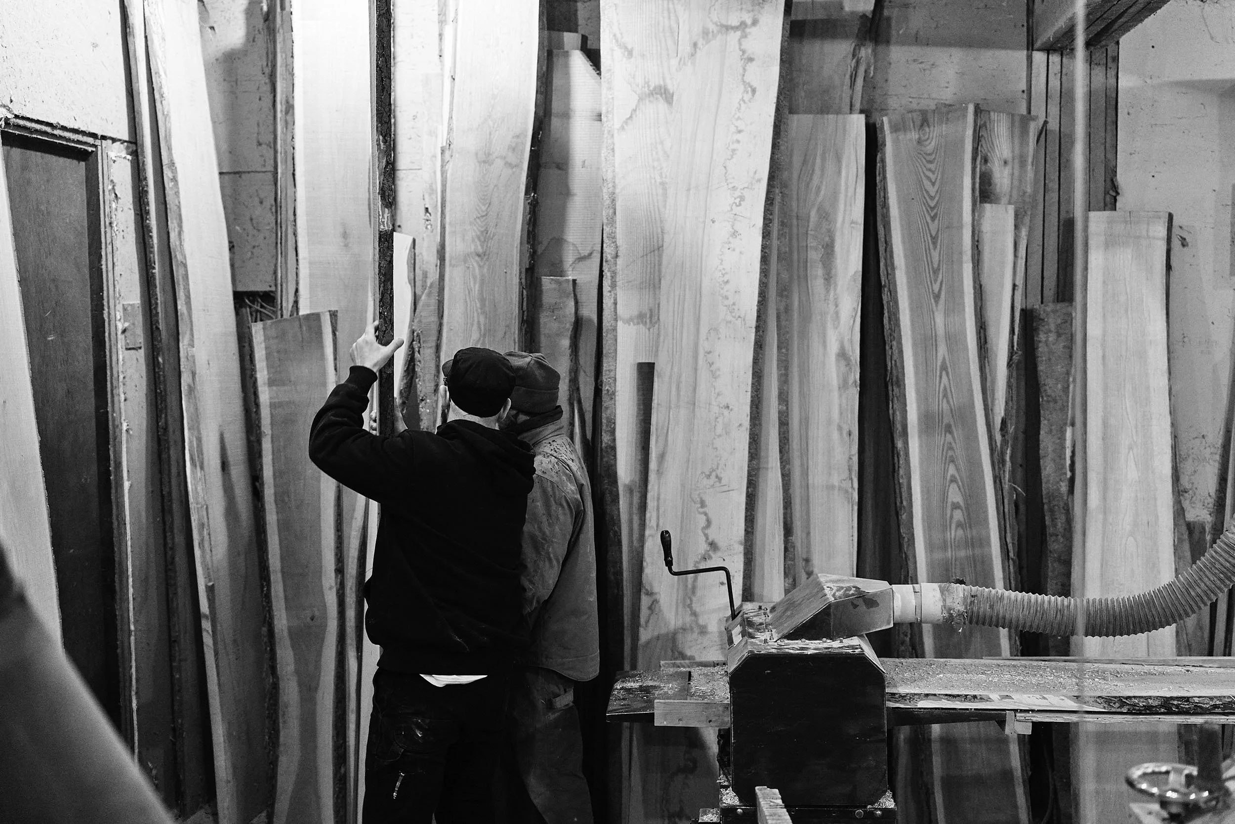A black and white image showing Aaron and Mark Speiser evaluate ten foot, live edge plank boards for use in lumberyard.  There are over 15 boards behind them as they evaluate one.