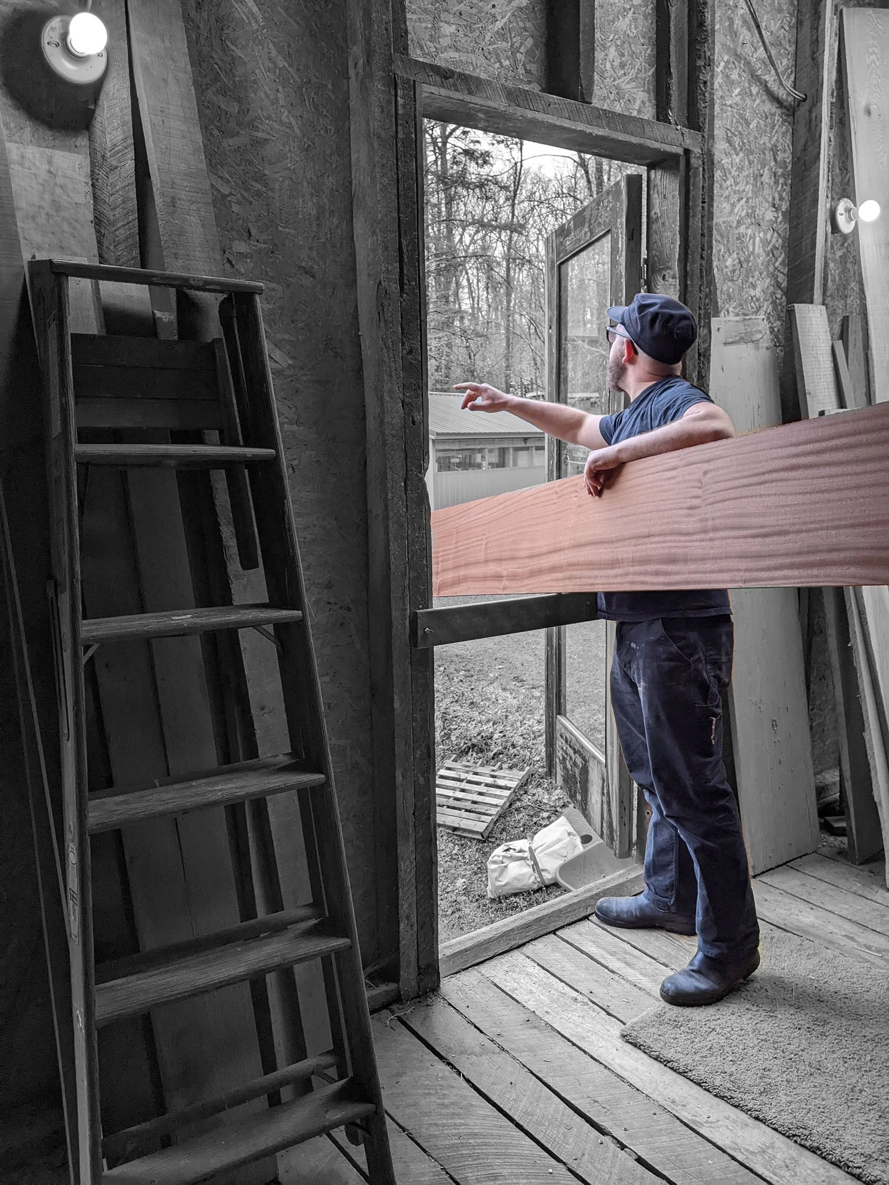 Aaron talks with Mark in the lumber loft through a window while holding a large piece of lumber through the window.