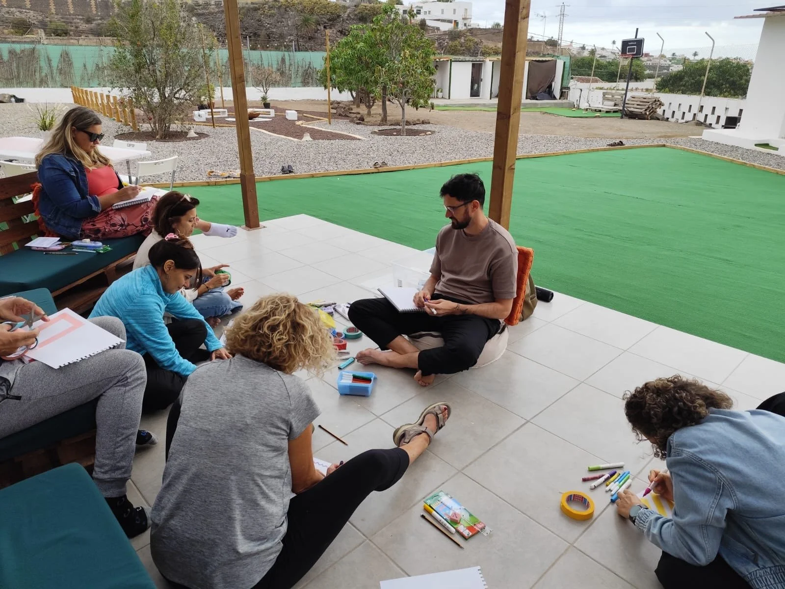 Grupo de personas participando en una sesión de entrenamiento o taller en un espacio abierto cubierto con vista a un área de césped y arboles, con materiales de papelería y notebooks.
