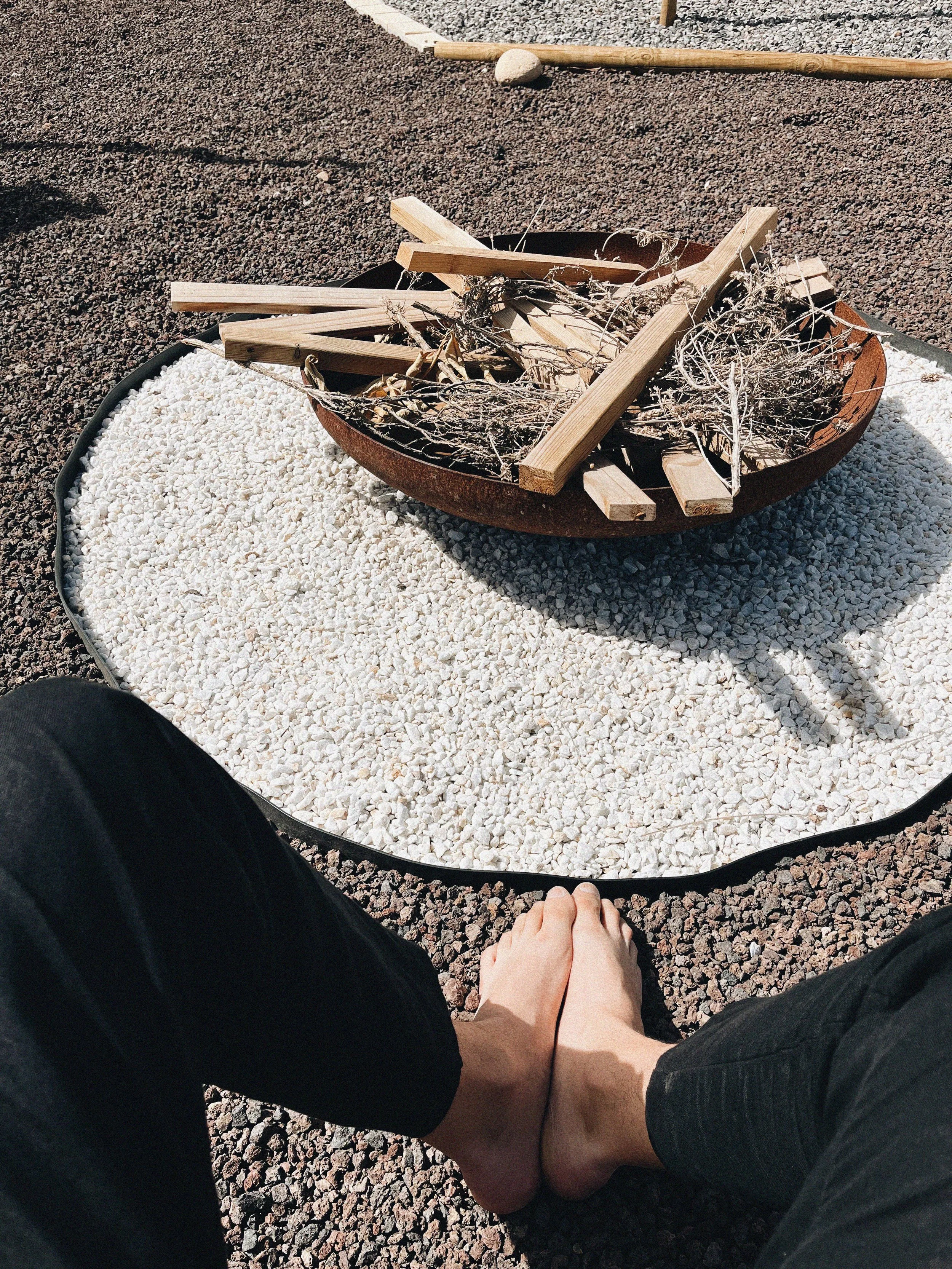 Pies descalzos delante de una zona decorativa con arena blanca y una fogata con madera seca y ramas secas.