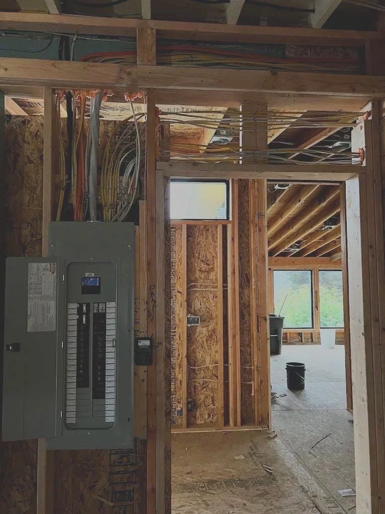 Interior view of a house under construction showing exposed wooden studs, electrical wiring, an electrical panel, and windows.