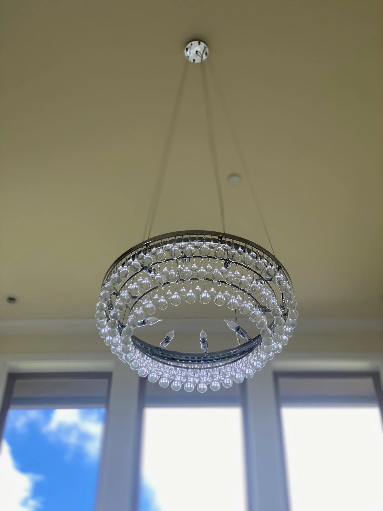 A chandelier with multiple tiers of clear glass or crystal globes hanging from a ceiling with large windows in the background.