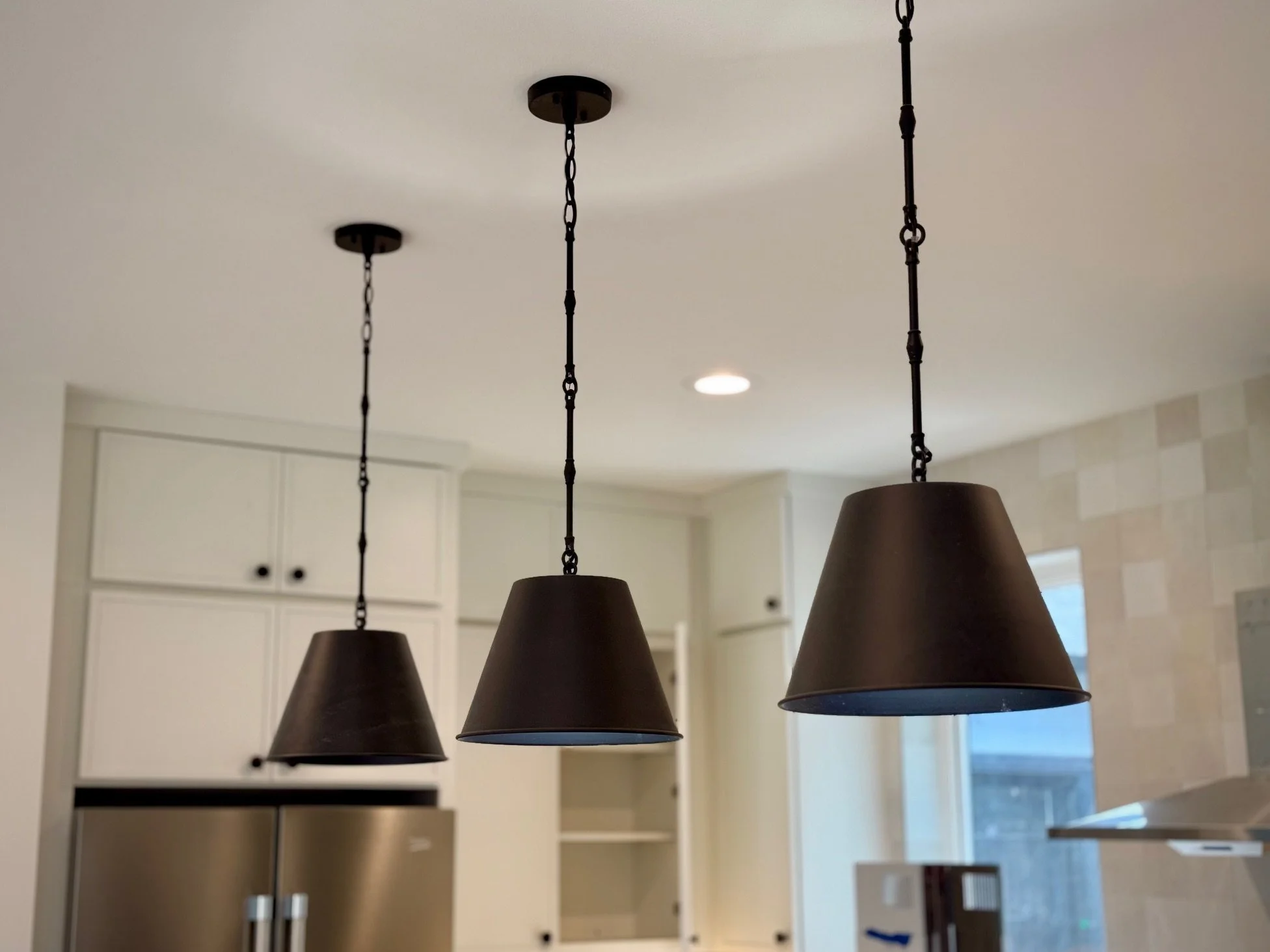 Three black pendant lights hanging from the ceiling in a modern kitchen with white cabinets and a stainless steel refrigerator.
