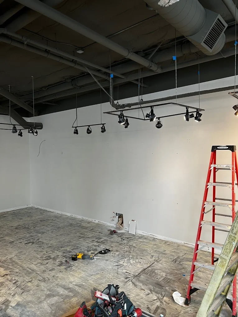 Room under renovation with unfinished flooring, a red step ladder, tools on the floor, and ceiling-mounted track lighting.