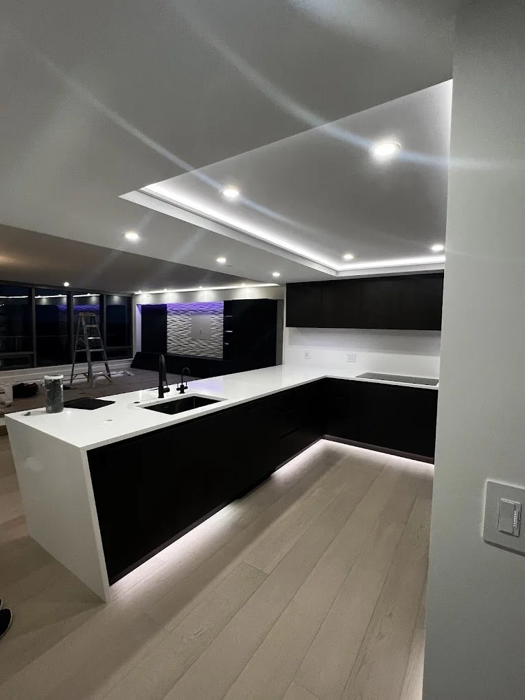 Modern kitchen with black and white cabinetry, white countertops, and recessed ceiling lighting. A ladder is visible in the background, and a view of a cityscape through large windows.
