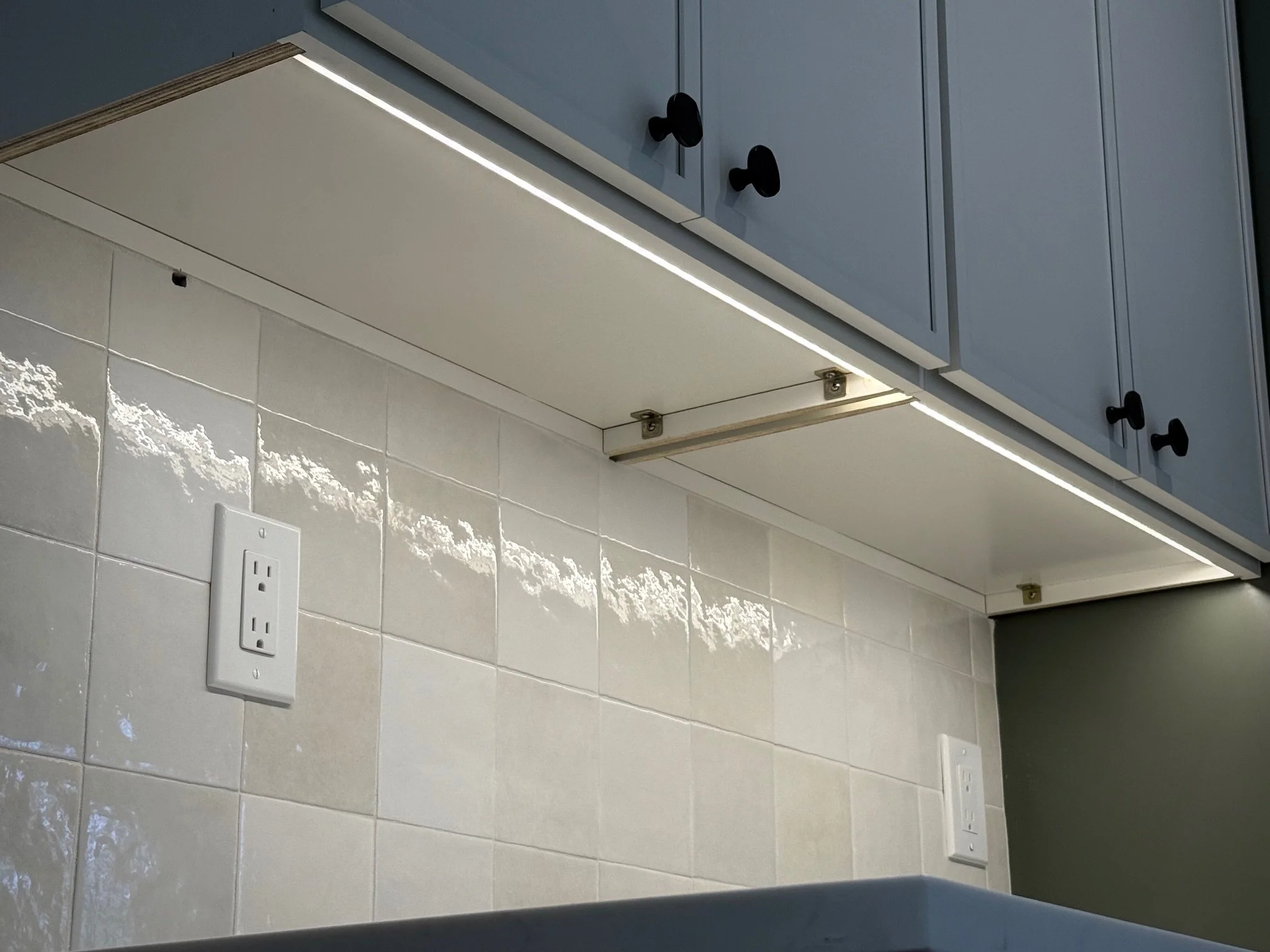 Under cabinet lighting illuminating a tiled kitchen wall with two electrical outlets.