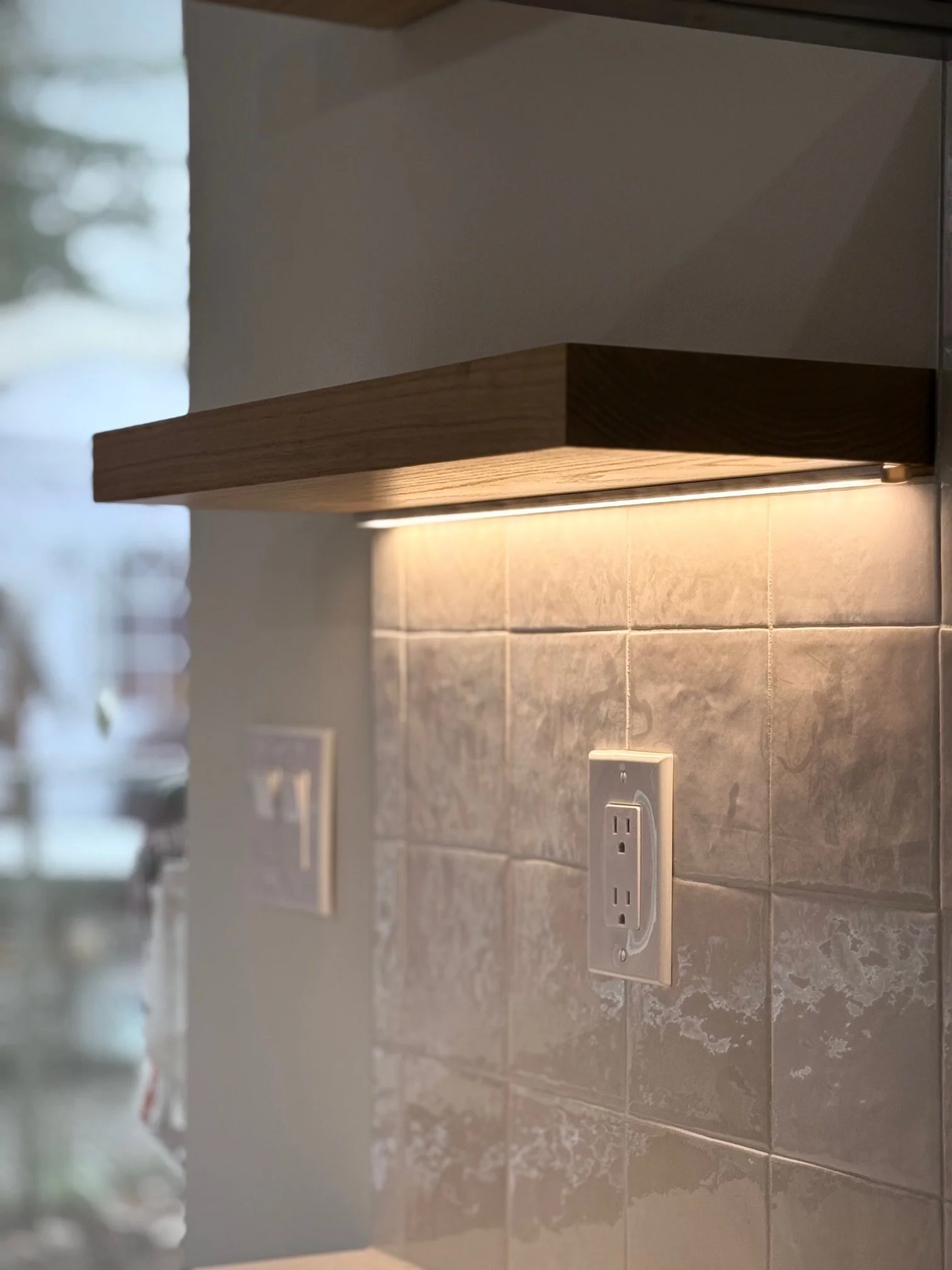Close-up of a kitchen wall with electrical outlets, tiled backsplash, a wooden shelf, and under-shelf lighting.