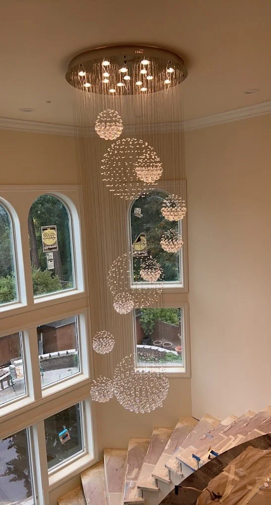 A multi-tiered crystal chandelier hanging in a room with large arched windows, seen from above, with a staircase in the foreground.