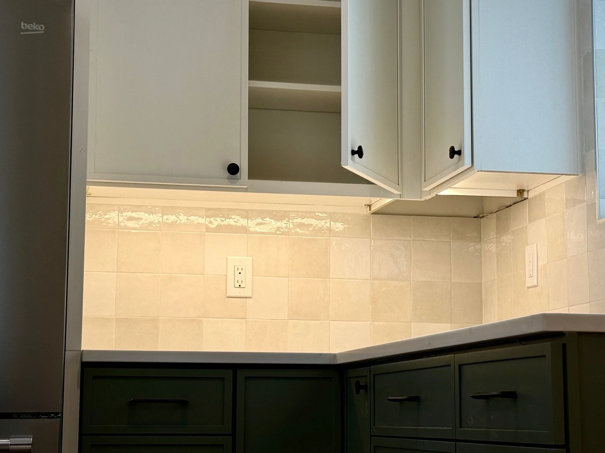 Kitchen with cream-colored tile backsplash, white upper cabinets, dark lower cabinets, and electrical outlets on the wall.