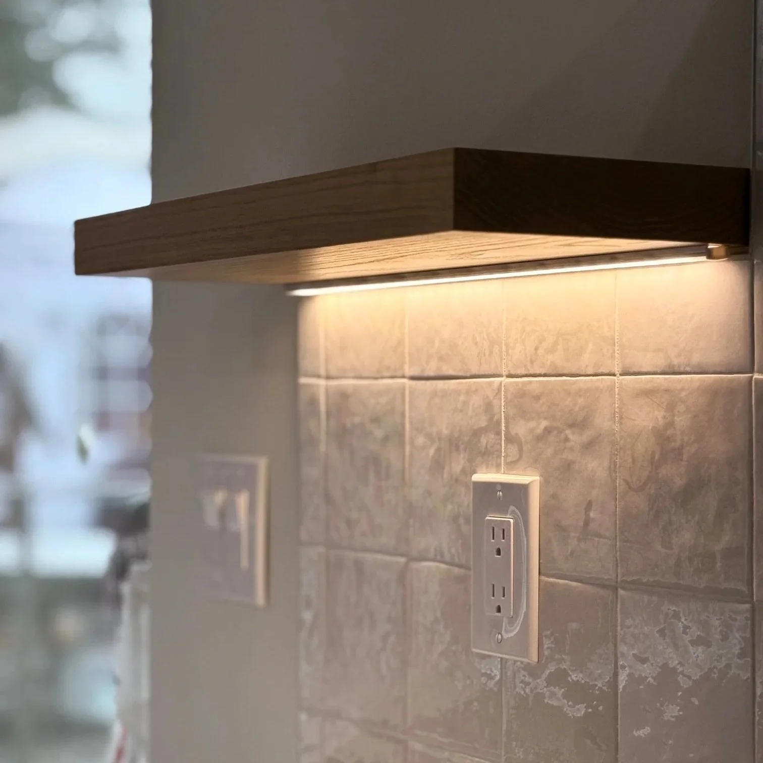 Close-up of a wooden kitchen shelf with built-in lighting, above a tiled wall with electrical outlets.