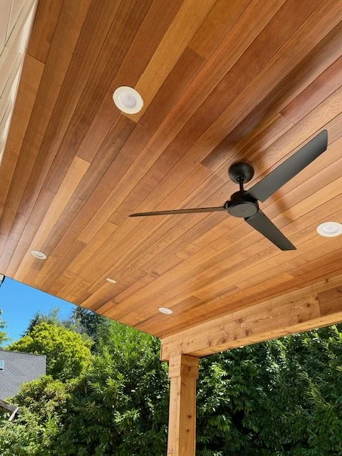 Wooden porch ceiling with recessed lights, a black ceiling fan, and green trees in the background.