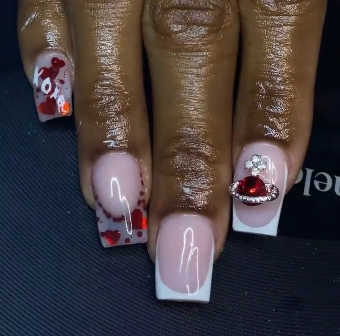 Nail art with pink base, red heart and love designs on the thumb and index finger, and a 3D red gemstone with rhinestones on the ring finger.