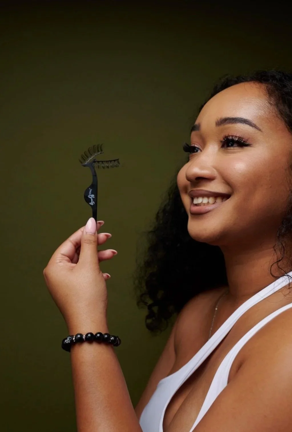 A woman with curly hair smiling, holding eyelash growth serum applicator in front of a green background.