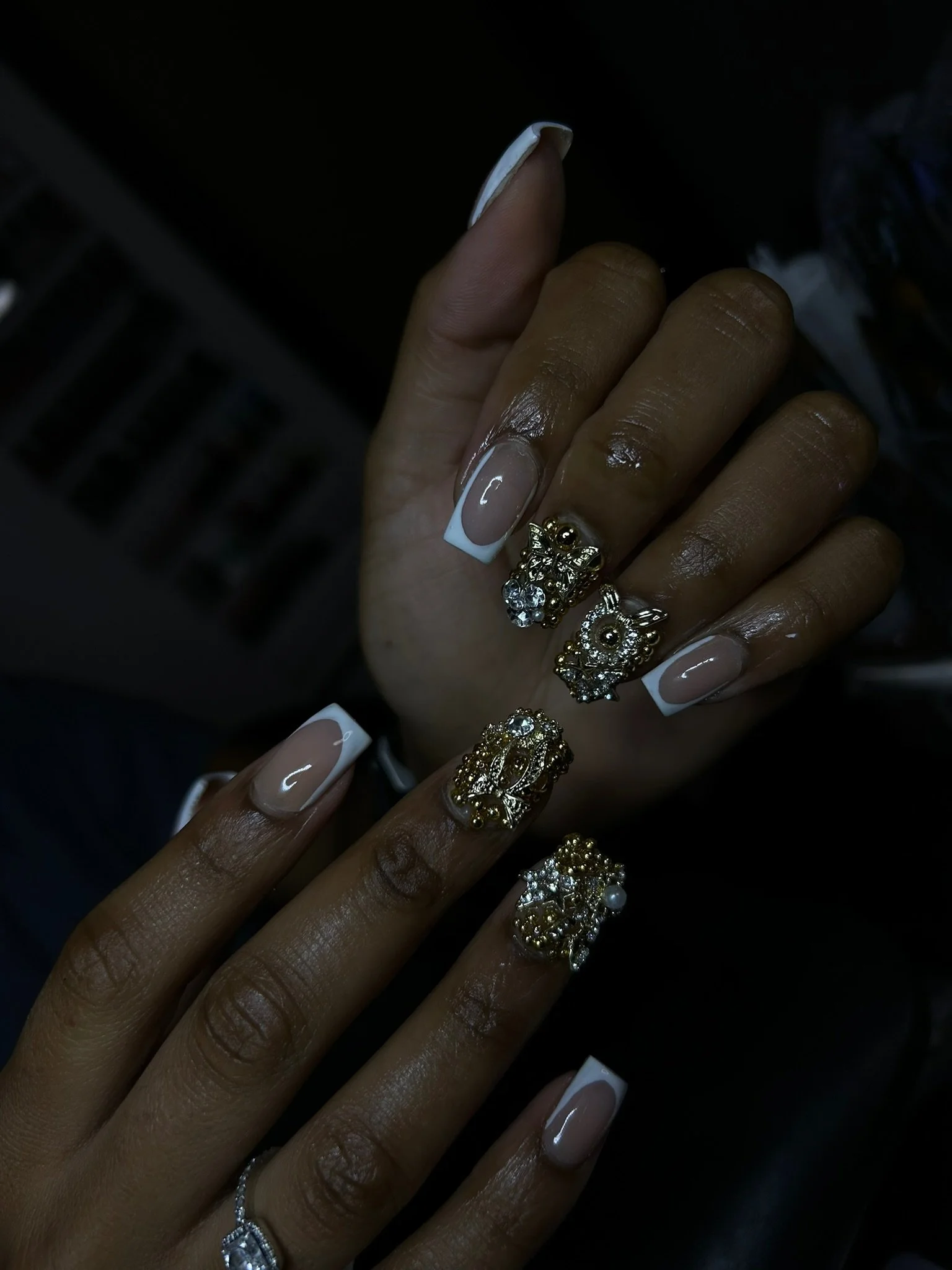 Close-up of hands with manicured nails and ornate owl-shaped rings, one hand with French manicure nails and the other with light pink nails, both decorated with shiny jewelry.