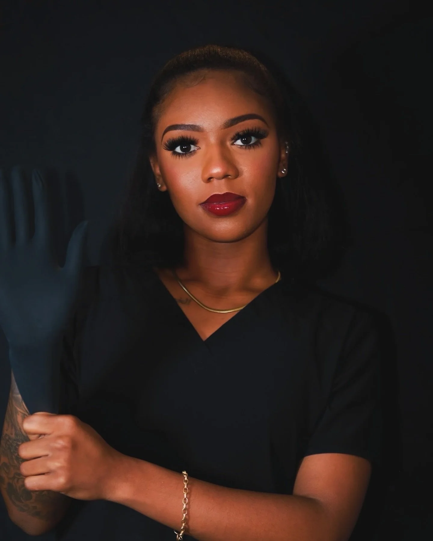 Portrait of a young woman with makeup, red lipstick, dark hair, wearing a black top, gold necklace and bracelet, posing against a dark background.