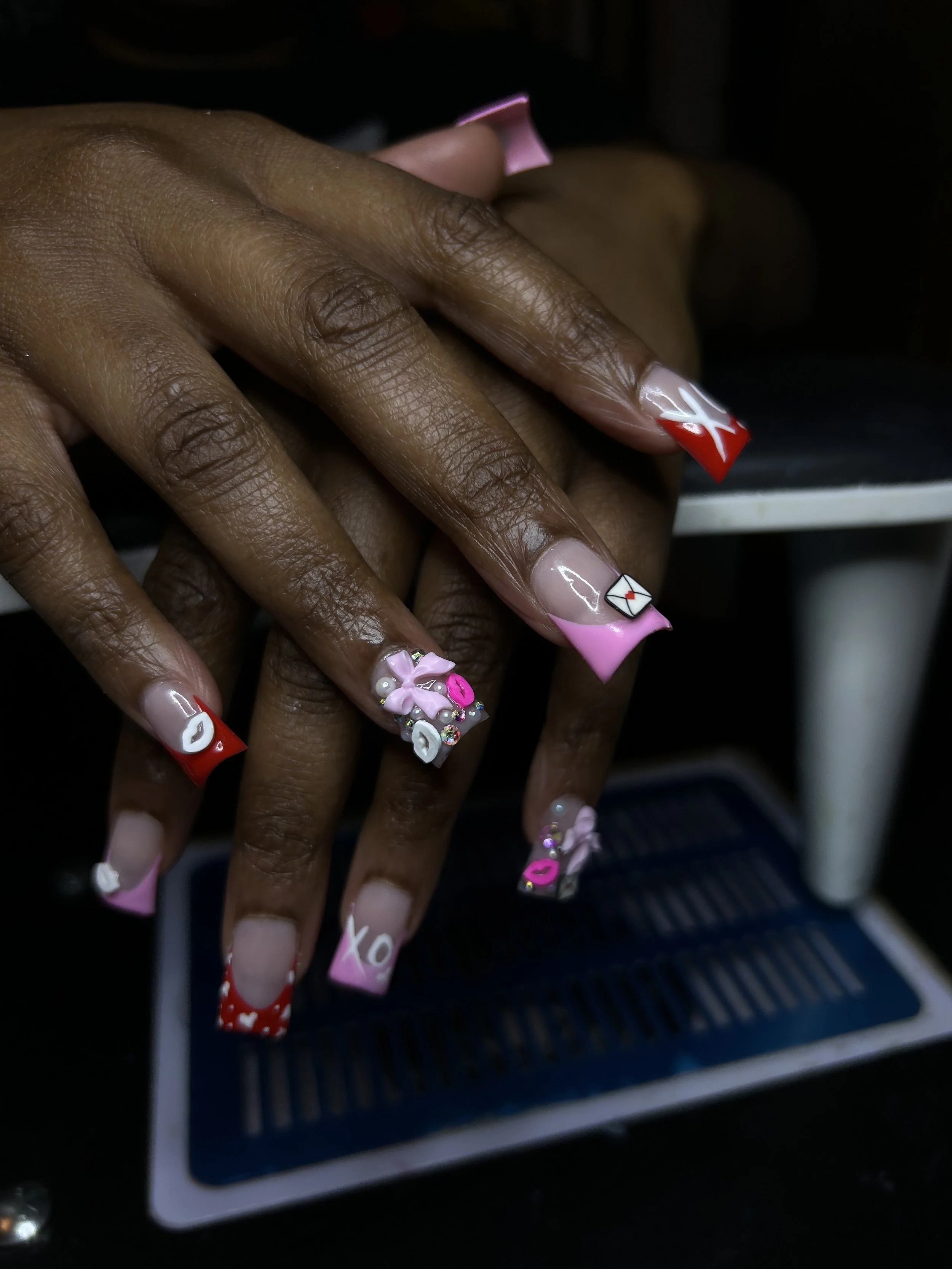 Close-up of a person's hands with decorated Valentine's Day-themed nail art, including hearts, lips, envelopes, and pink 3D flowers, with a dark background.