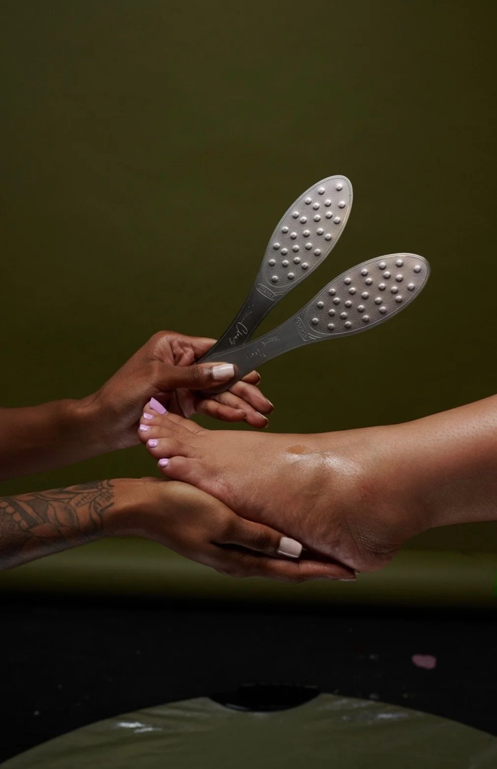 Two hands holding three metal foot files, with one foot hand resting on a black surface and the other on a person's lap, against a dark green background.