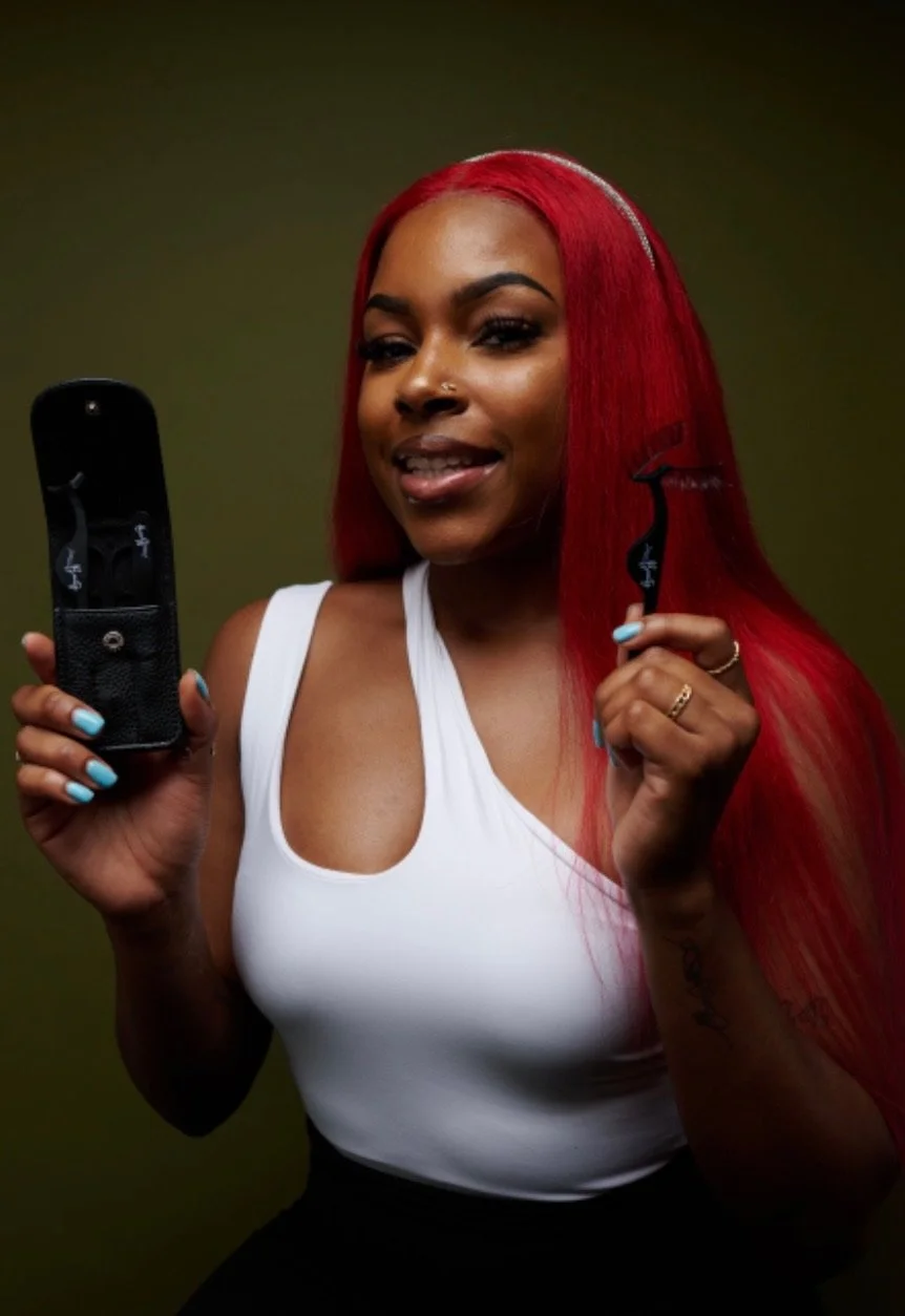 A woman with bright red hair, wearing a white sleeveless top, holding a curling iron in one hand and a curling iron clip in the other, smiling at the camera.