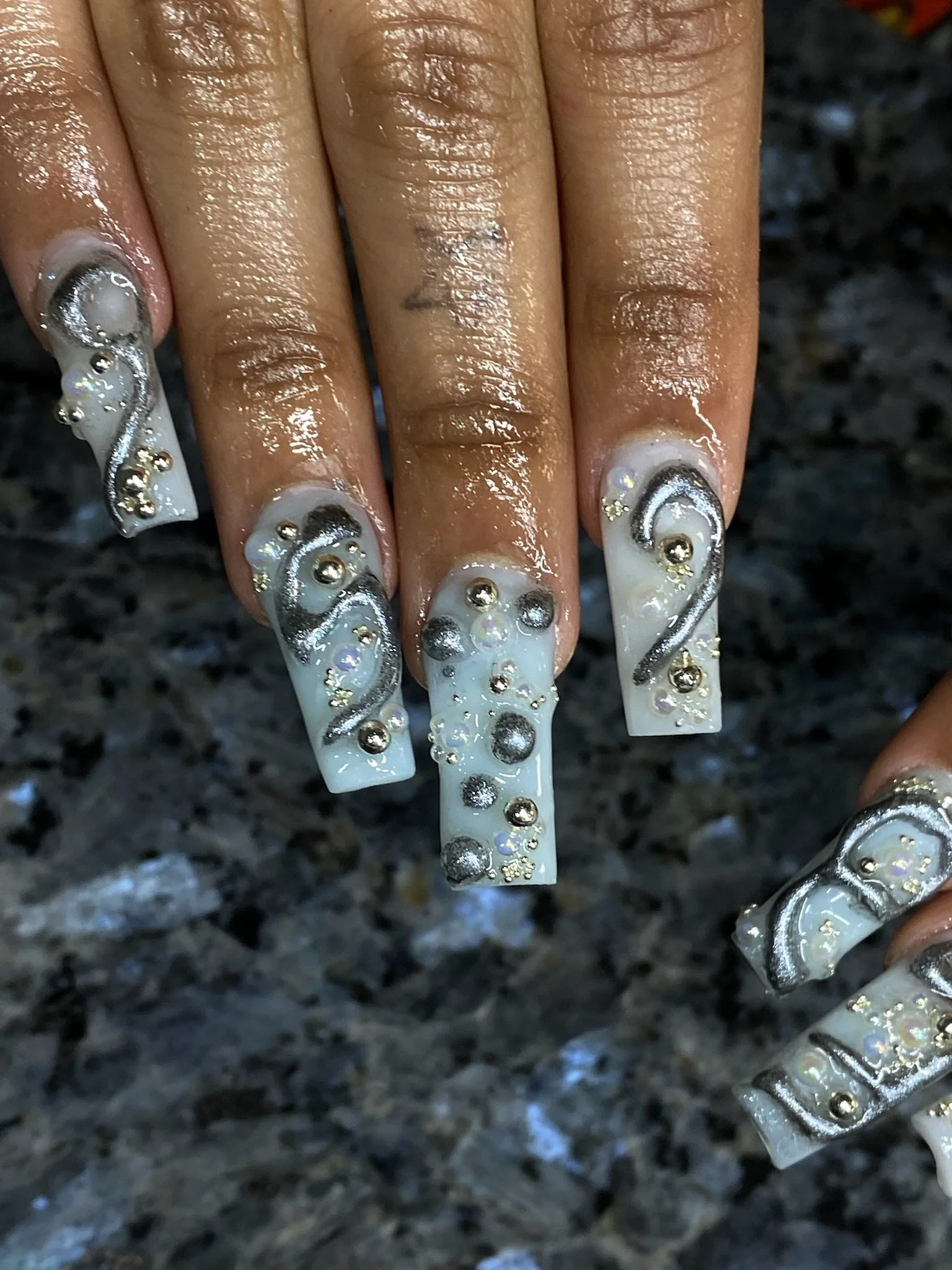 Close-up of a person's hand with long, decorated nails featuring white and silver designs, rhinestones, and 3D embellishments, on a dark, speckled surface.