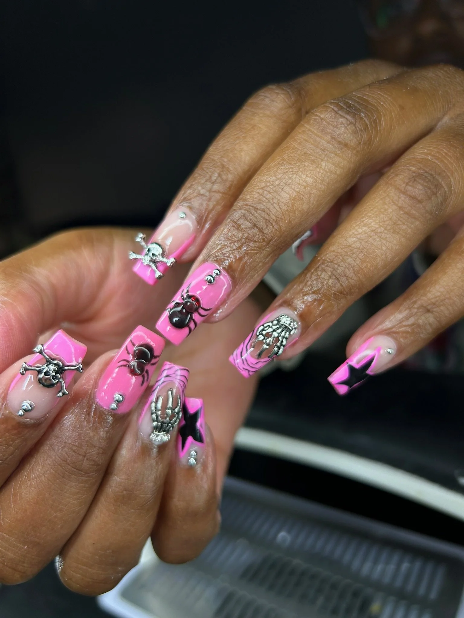 Close-up of a hand with decorated pink nails, featuring skull and crossbones charms, black stars, spider web patterns, and small rhinestones.