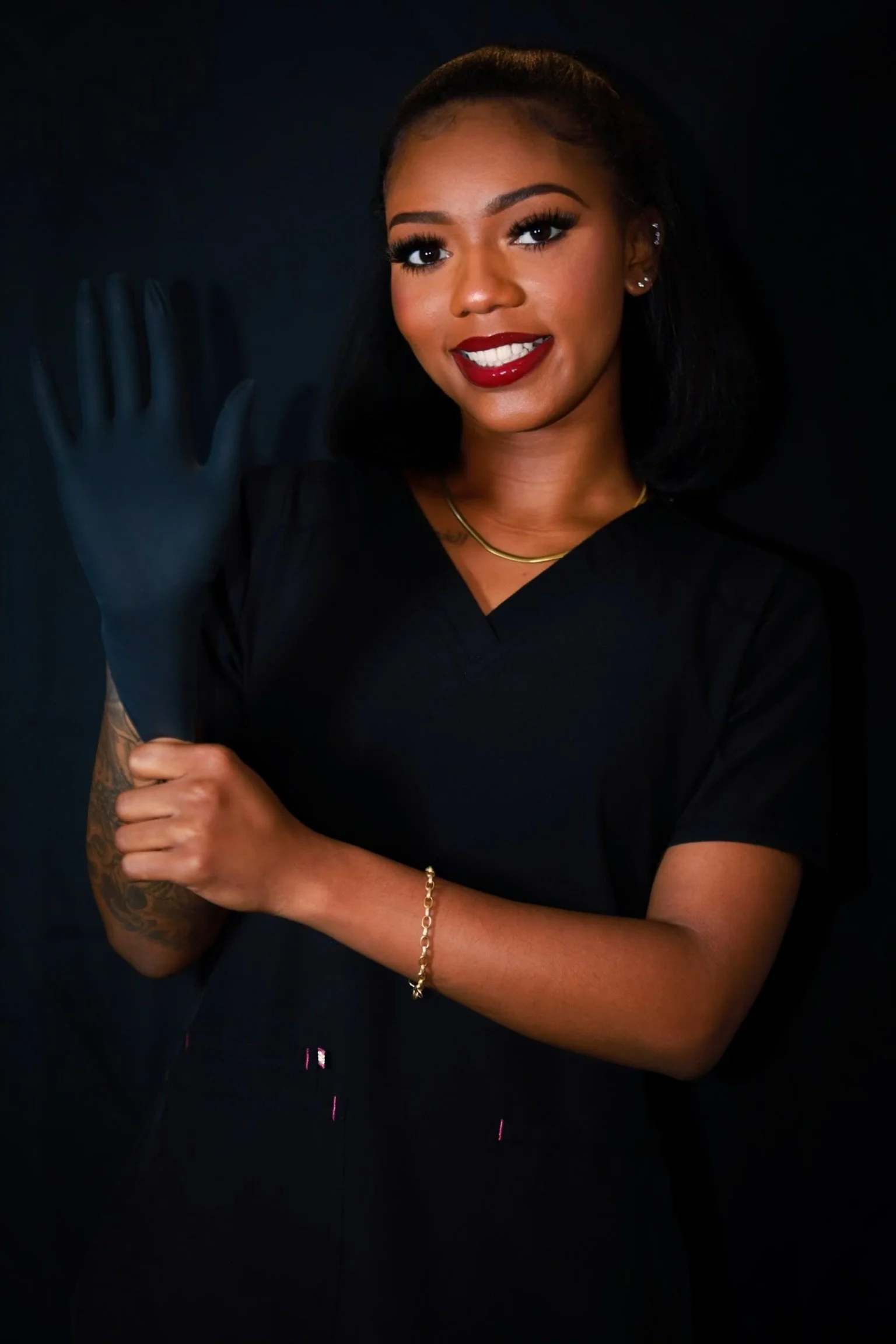 A woman with dark hair, wearing a black uniform, red lipstick, and a gold necklace and bracelet, smiling and holding up a black glove.