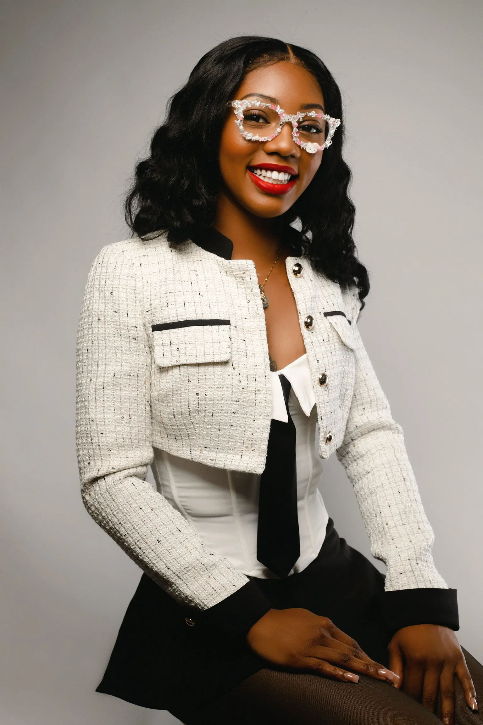 A woman smiling, wearing colorful glasses, a white textured jacket, a white blouse, a black tie, and black skirt, sitting against a plain background.