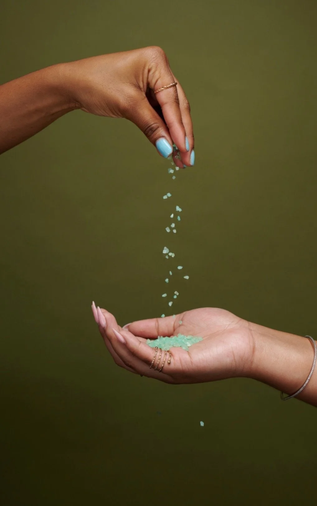 One person's hand is dropping small teal-colored salt crystals into another person's open hand against a solid green background.
