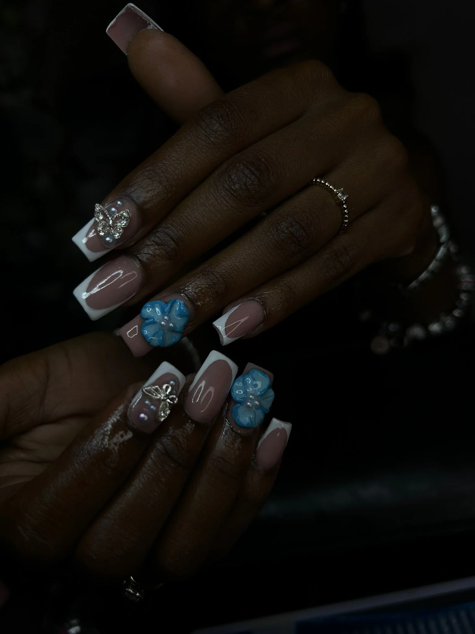 Close-up of a person's hand and nails with detailed nail art, featuring French tips, 3D blue flowers, pearl accents, and silver jewelry including rings and a bracelet.