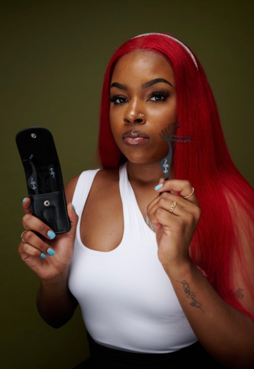 Woman with long red hair holding a hair cutting tool in one hand and a pair of scissors in the other, dressed in a white top, with a confident expression.