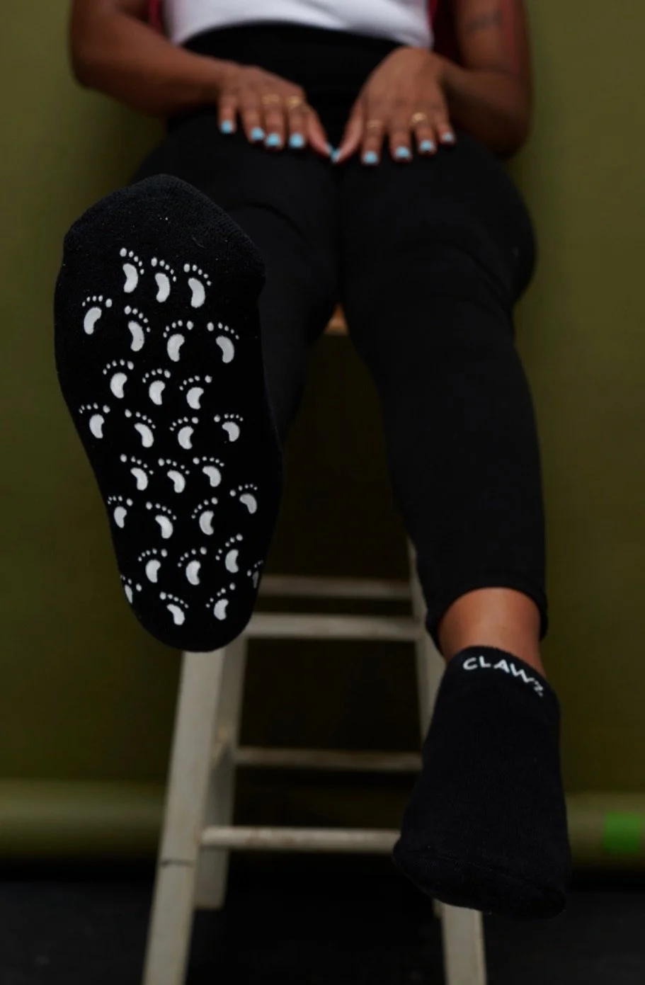 A person sitting on a stool with their legs raised, showing the sole of a black sock with white dotted crescent shapes and the word 'CLAWS' on the ankle.