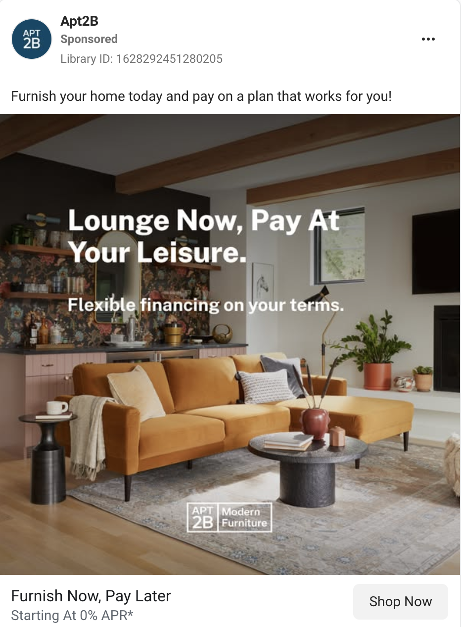 A modern living room with a mustard-colored sofa, a round black coffee table, and decorative plants. There is a window with natural light, a wall-mounted TV, and open shelves with books and decor.