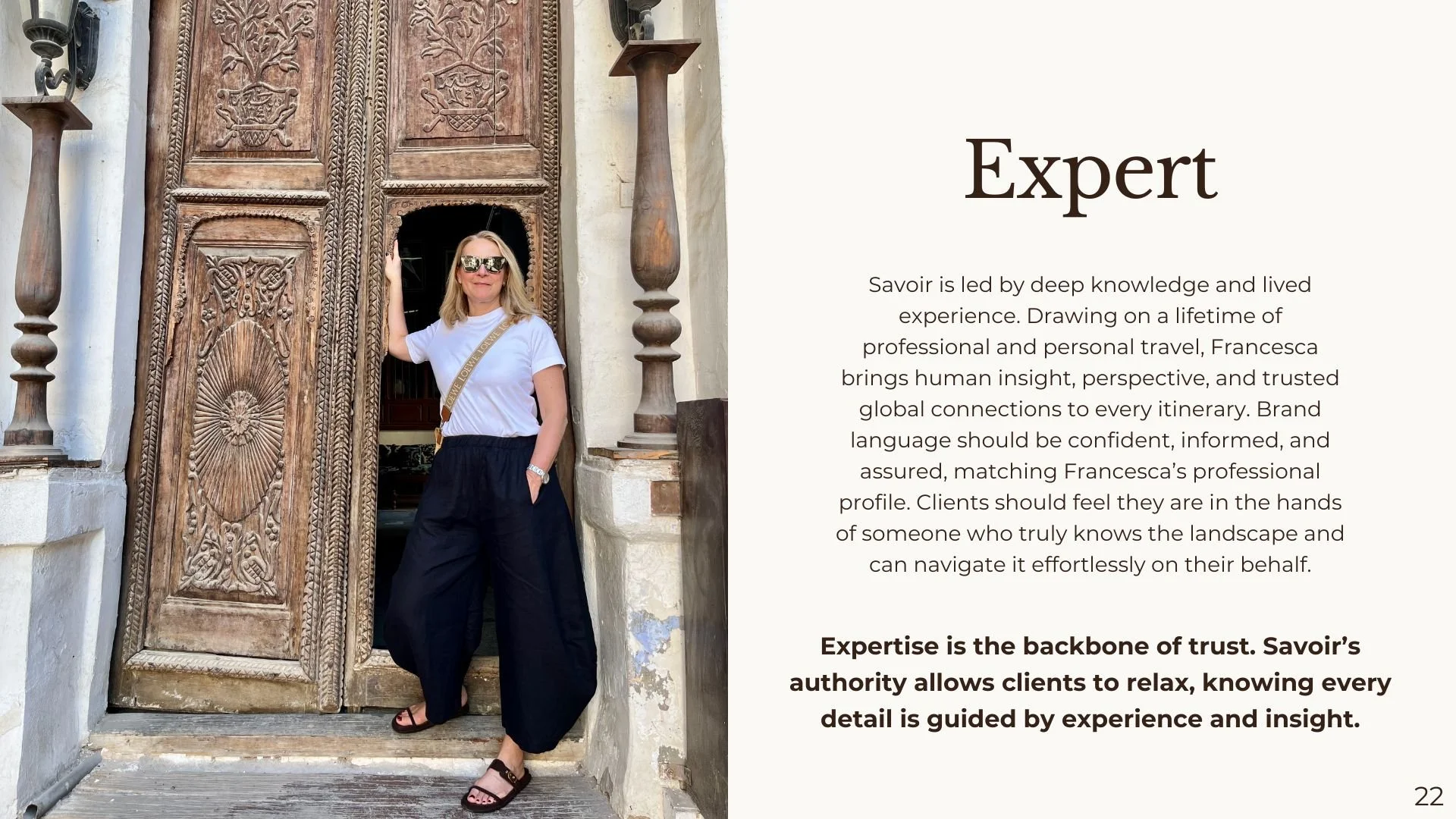 A woman wearing sunglasses, a white t-shirt, and black pants stands in a doorway with intricately carved wooden doors. She is smiling and resting her hand on the door frame, carrying a beige crossbody bag and wearing a watch.