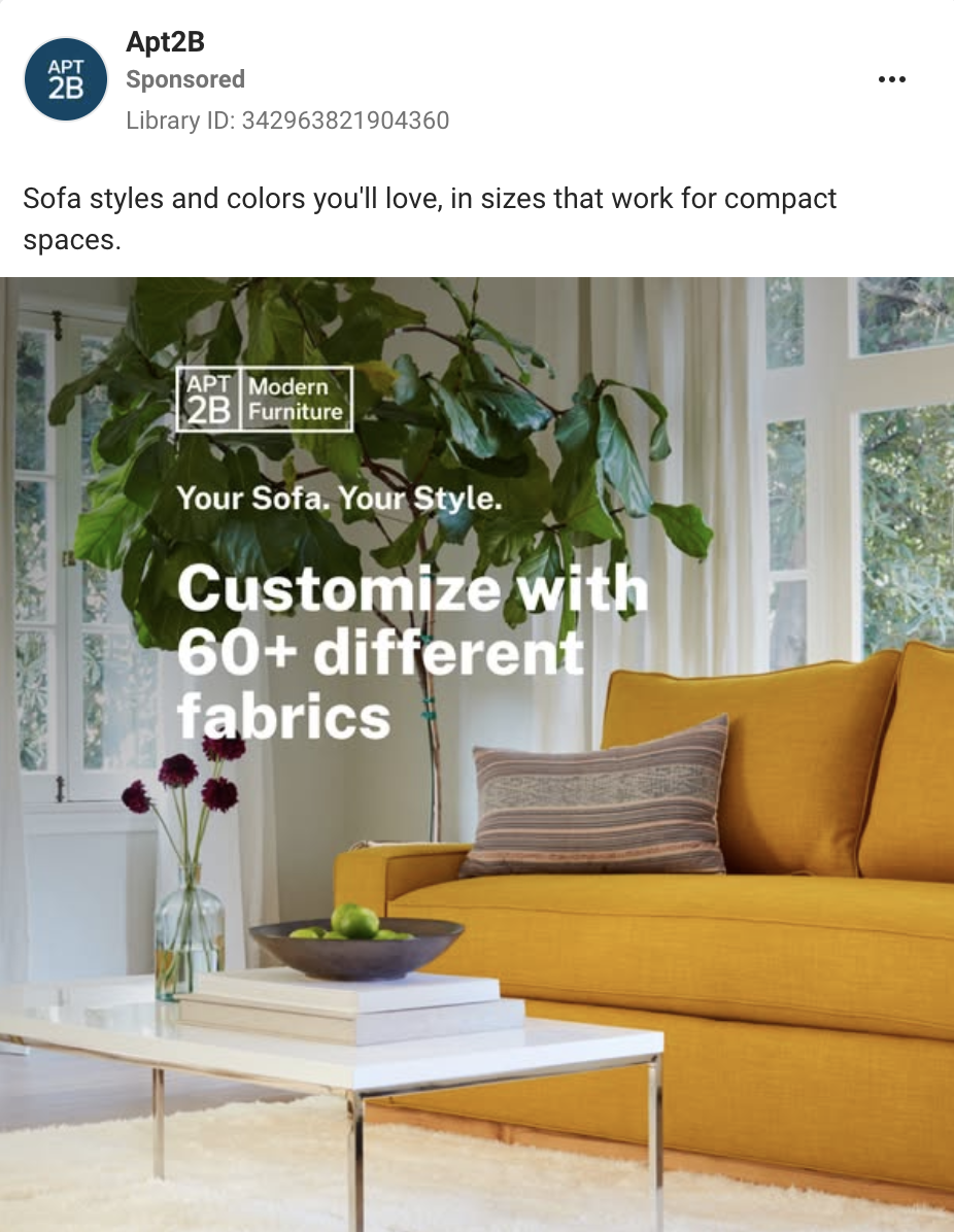 Interior of a living room with a yellow sofa, a striped pillow, a white coffee table with a bowl of limes and a stack of books, a vase with flowers, large windows, and a tall plant.