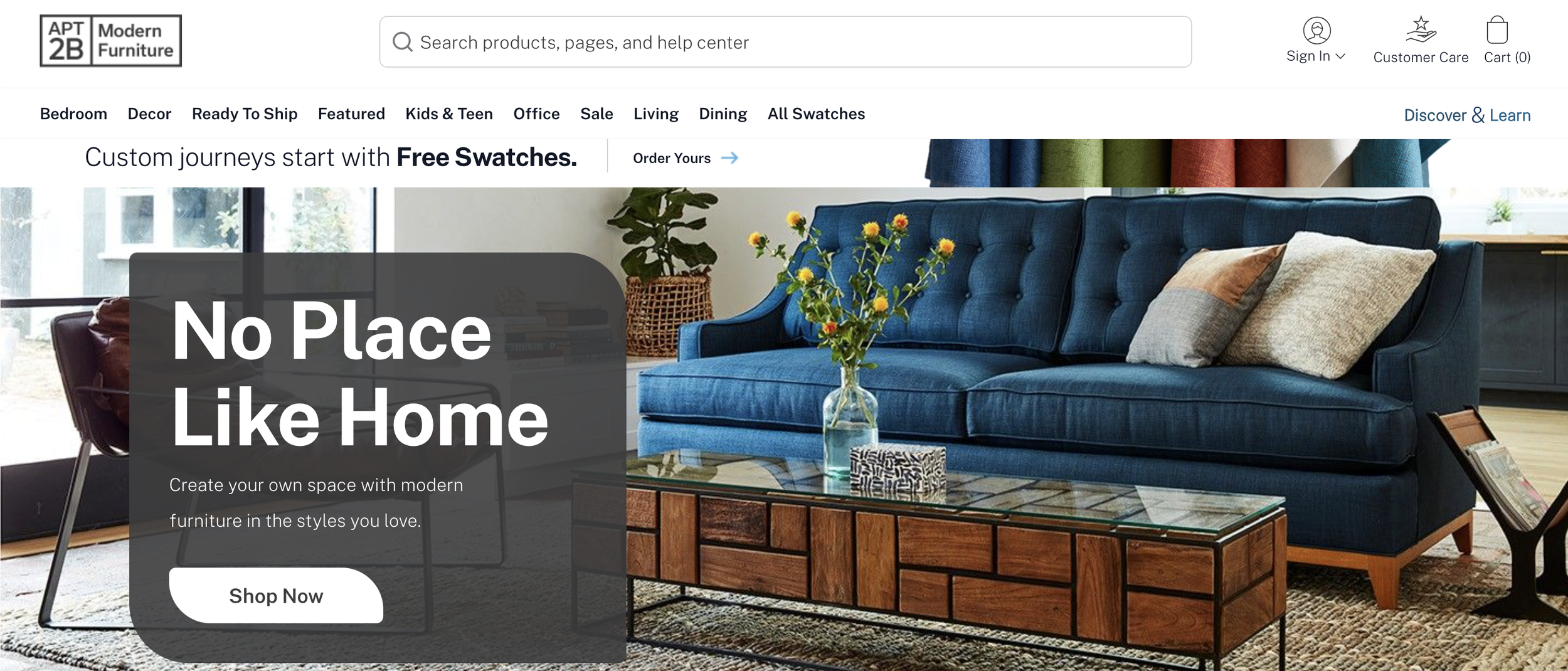A modern living room with a bright blue sofa decorated with beige and brown pillows, a glass coffee table, a glass vase with yellow flowers, and a basket on a side table. The room has large windows with sunlight coming in.