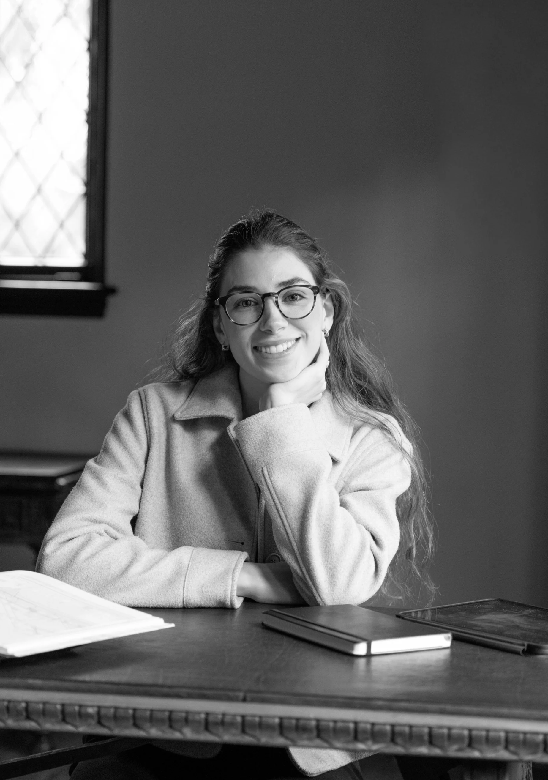 A smiling young woman with glasses, long curly hair, wearing a light-colored coat, sitting at a wooden desk with a book and a tablet.