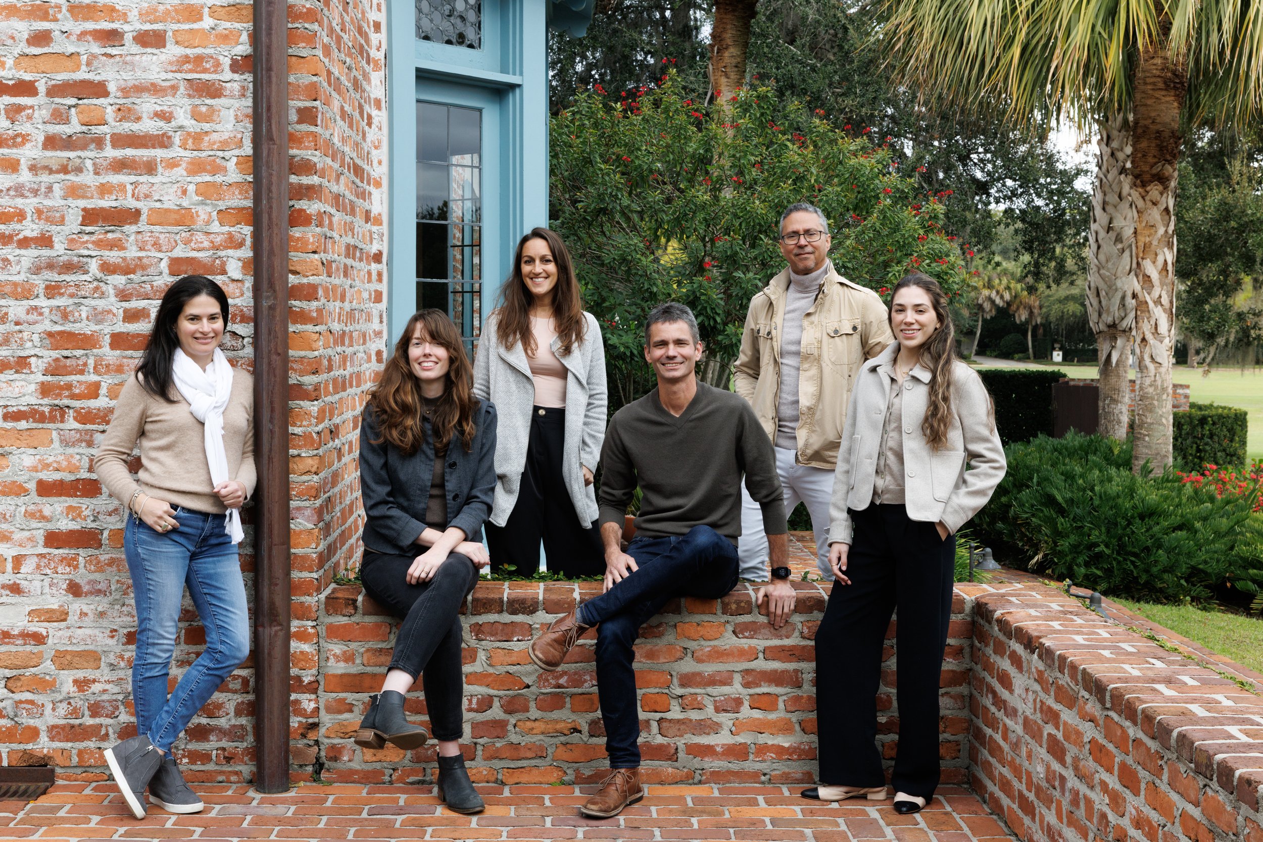 Group of seven people outdoors, standing and sitting on a brick ledge in front of a brick building with a blue window frame, surrounded by green trees and plants.