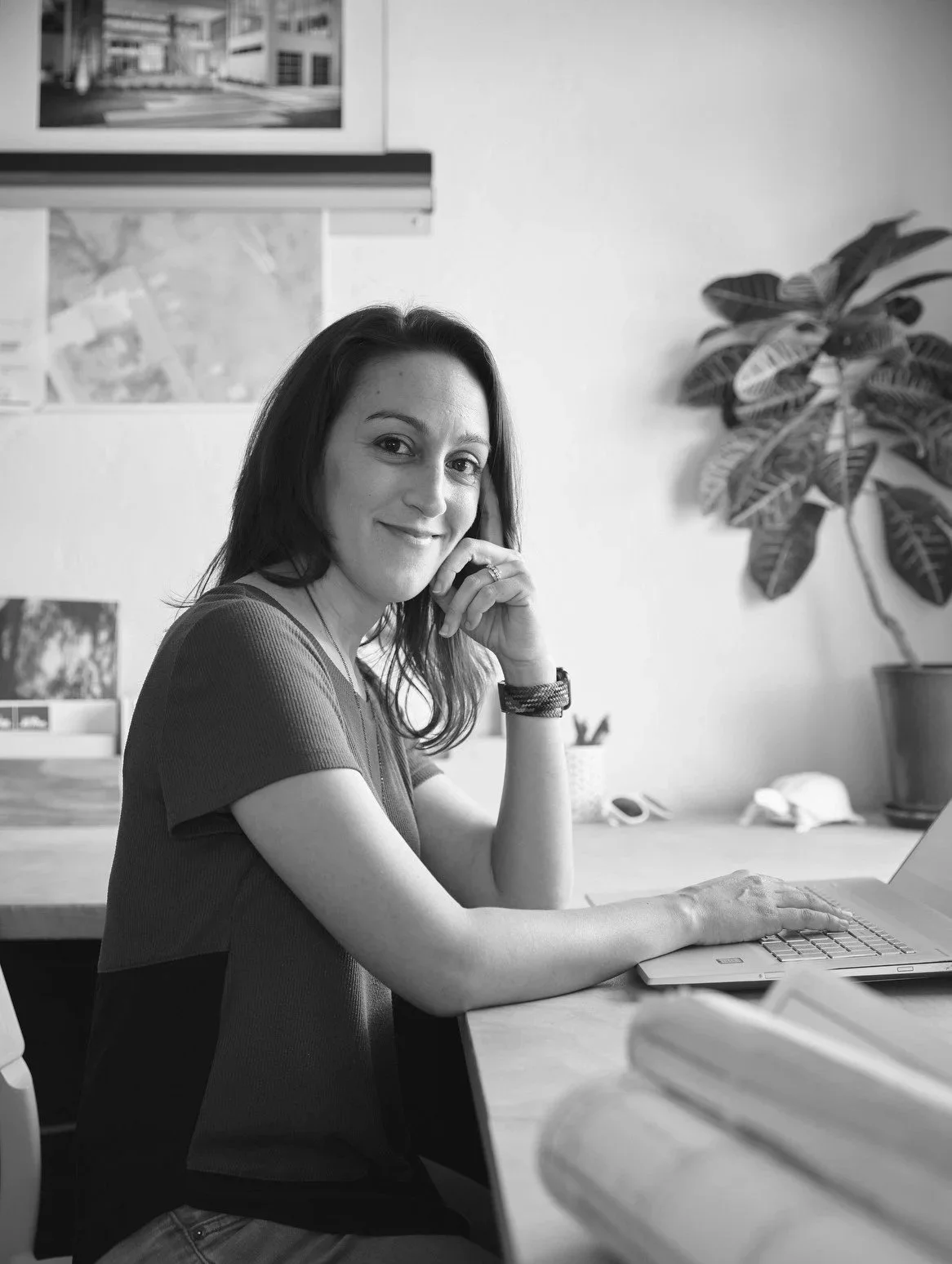 A woman sitting at a desk in an office, smiling at the camera, with a laptop in front of her and a large potted plant in the background.