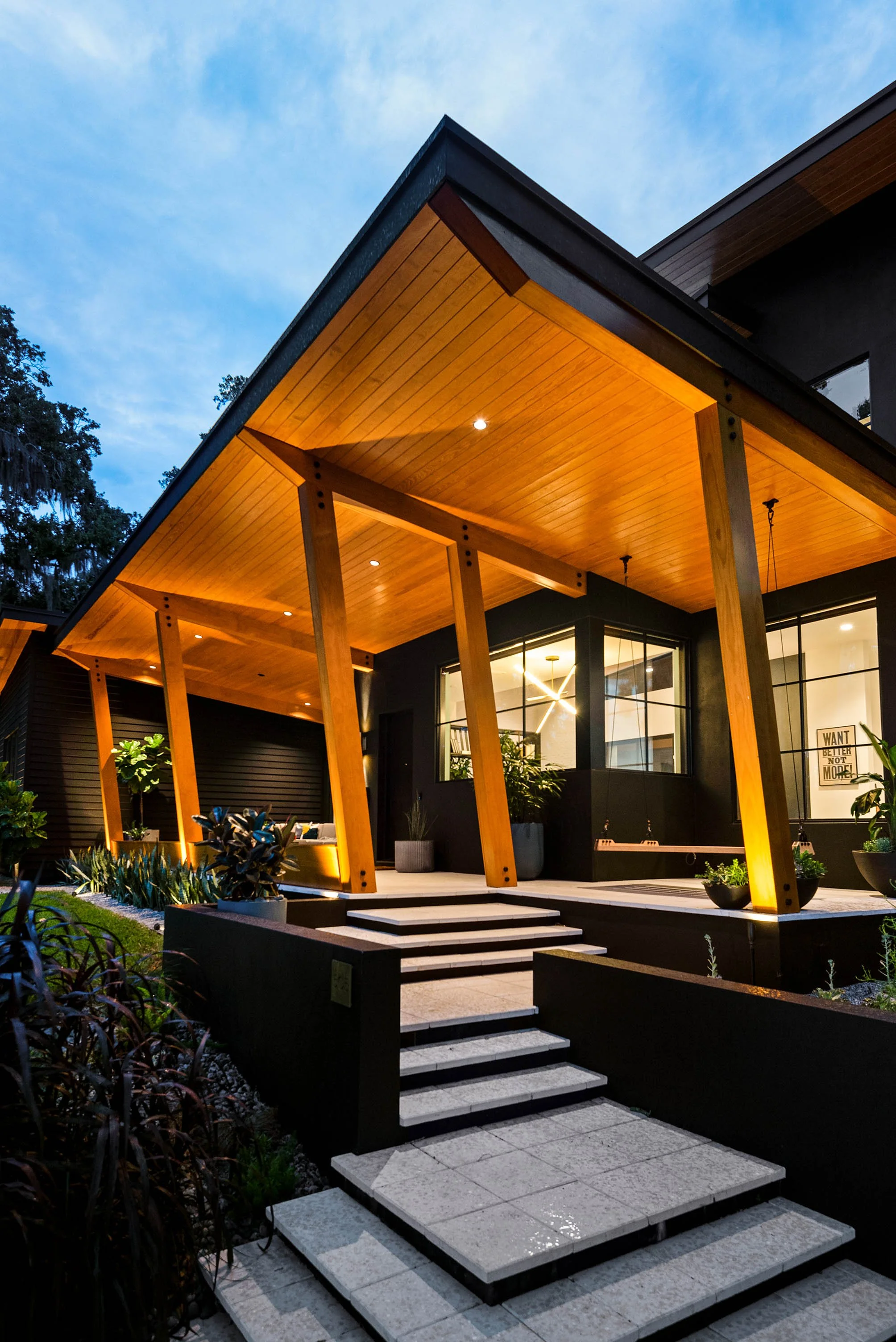 Modern house with wooden porch, outdoor lighting, plants, and large windows at dusk